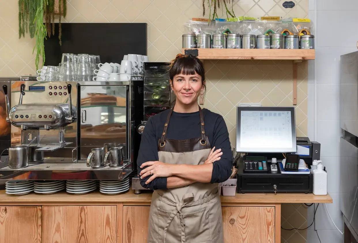 A working woman in a café business, standing behind the counter next to the cash register, with arms crossed, looking at the camera.